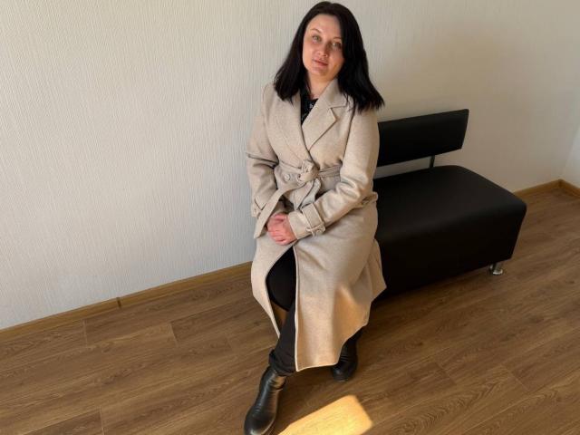 LinaSpring, una donna dalla pelle chiara con capelli neri, è seduta su una panchina nera, indossando un cappotto beige e pantaloni neri, con le gambe leggermente distanziati, con il pene visibile, pavimento in legno, parete bianca.