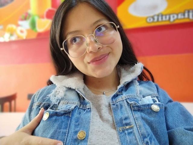 AnittaRouses with long black hair, wearing gold-rimmed glasses and a frayed denim jacket over a white shirt, smiling slightly. Background features bright red and orange colors. Casual, confident pose.