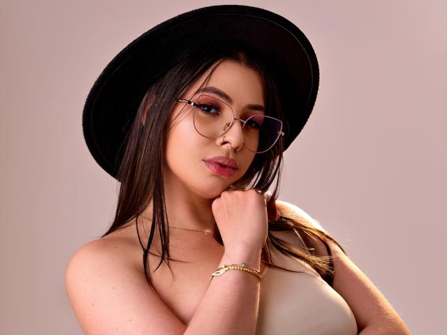 Photograph of VanessaClose, sexy, wearing a black hat, round glasses, and a gold bracelet, looking seductively at the camera with a hand near her face, wet black hair, white top.