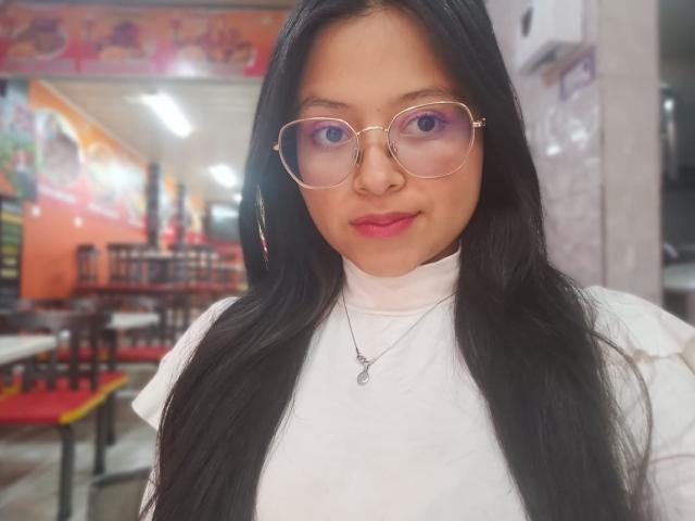 AnittaRouses with long black hair, wearing glasses and a white turtleneck, standing in a brightly lit indoor setting with red and orange decorations. Background includes chairs and tables. She has a subtle, confident smile.