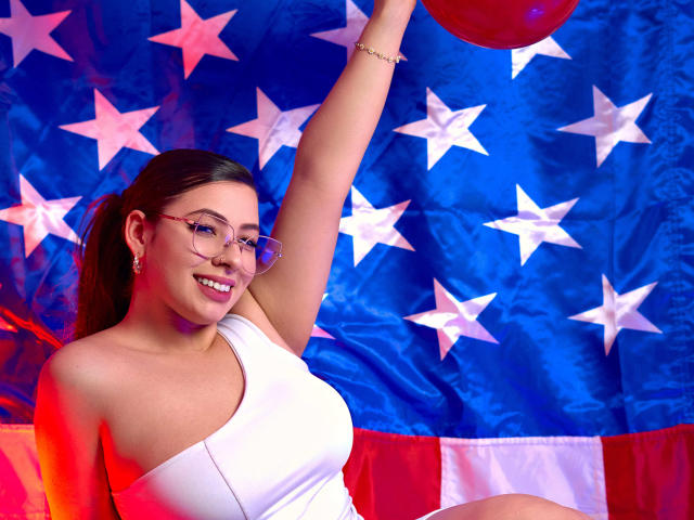 VanessaClose, Asian woman with long black hair, wearing glasses and a white one-shoulder top, raising her right arm, against a vibrant American flag backdrop, smiling confidently. Bright colors, bold stars.