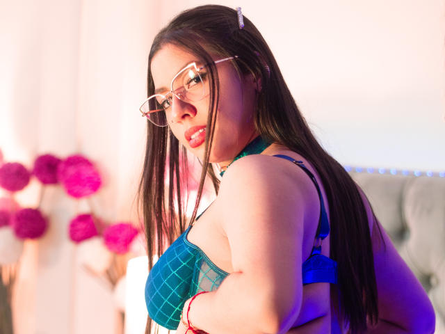 Photograph of VanessaClose, a Latina woman with long black hair, wearing glasses and a blue, patterned bra, looking back over her shoulder with a seductive expression. Vibrant pink flowers in background.