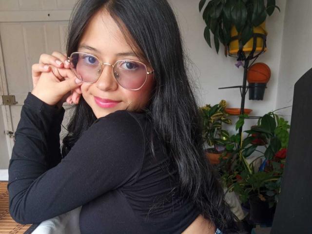 AnittaRouses, an Asian woman with long black hair, wearing a black long-sleeve top, adjusting gold-rimmed glasses with her right hand, blue eyes, slightly smiling, surrounded by potted plants.
