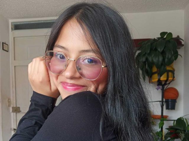 Photograph of AnittaRouses with long black hair, wearing round gold glasses, black top, and pink lipstick, touching glasses with right hand, blue eyes, plant in background, slight smile. Fucking cute.