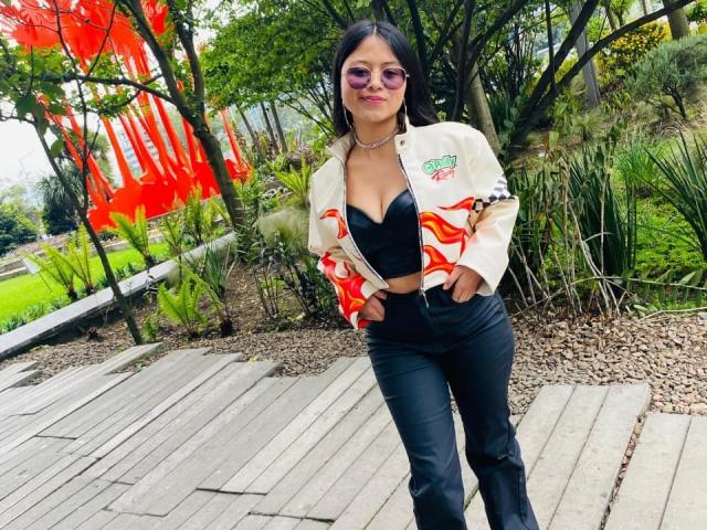 AnittaRouses stands on a wooden path, wearing a white jacket with red accents, black pants, and a low-cut black top revealing cleavage. She's looking at the camera with sunglasses. Background includes trees and red fabric.