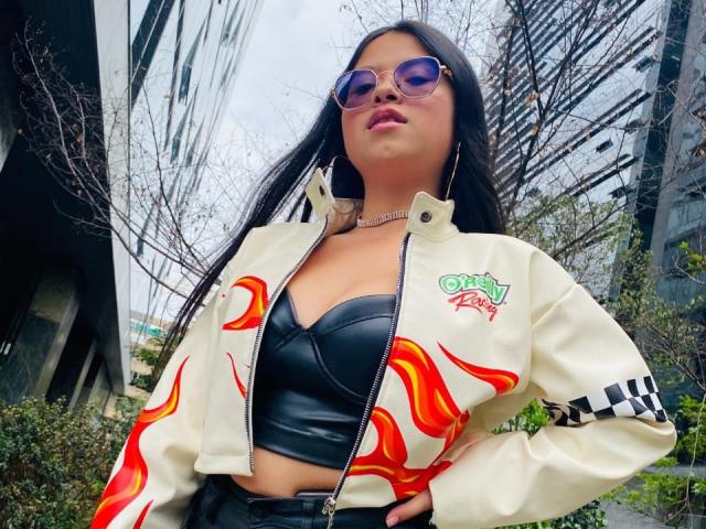 Anitta trägt langes, schwarzes Haar, eine Sonnenbrille, eine weiße Jacke mit roten Flammen, ein schwarzes Lederoberteil und eine Halskette. Städtische Hintergrundkulisse, Pflanzen an der Straße. Selbstbewusste Pose, verführerische Ausstrahlung..