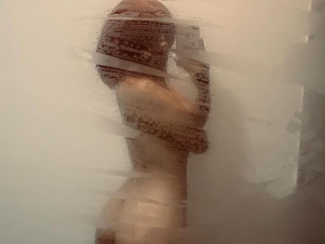 Photographie floue d'une BelladonnaLips nue, avec la peau mouillée, debout, les bras croisés sur la poitrine, créant une ambiance brumeuse. Effet de vapeur. Pénis érigé visible, tension sexuelle, pose suggestive.