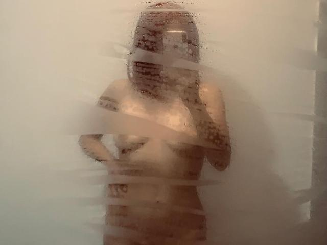 Vervormde foto van BelladonnaLips die in een douche staat, met waterdruppels op het glas, waarbij ze hun borsten vasthouden, gedeeltelijk. zichtbare, opgewekte tepels, dampig en mistig, seksuele spanning, natte huid, erotisch.