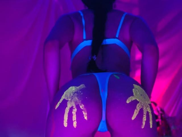 BelladonnaLips está arrodillada sobre una cama, de espaldas a la cámara, vistiendo una tanga y un sujetador azules. Huellas brillantes cubren su trasero. Iluminación de color rosa neón y azul. Muy atractivo/a..