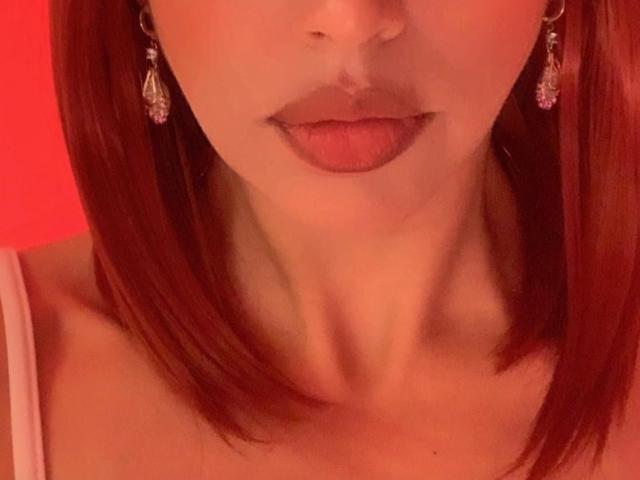 Gros plan du visage de BelladonnaLips, éclairé d'une lumière rouge. Elle a les cheveux roux, porte des boucles d'oreilles pendantes, et une bretelle blanche est visible sur son épaule. Ses lèvres sont légèrement entrouvertes, laissant entrevoir une certaine tension..