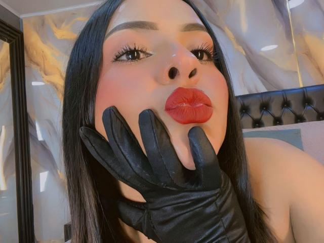 Nahaufnahme von LindaFranco mit langen, schwarzen Haaren, roten Lippen und schwarzen Lederhandschuhen, wobei eine Hand verführerisch das Gesicht bedeckt. Glänzende Augen, blasse Haut, ein schwarzer, mit Fransen versehener Kopfteil im Hintergrund. Verdammt heiß..