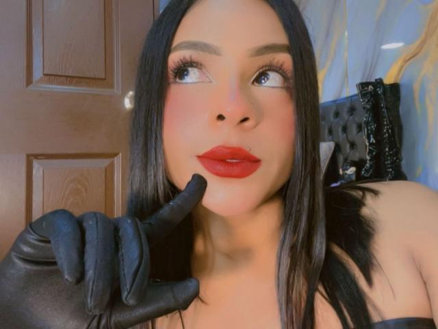 Primer plano de LindaFrancoo con cabello negro, lápiz labial rojo y guantes negros, con un dedo tocando sus labios, mirando hacia arriba. Expresión sensual. El fondo incluye una puerta de madera y cuero negro..