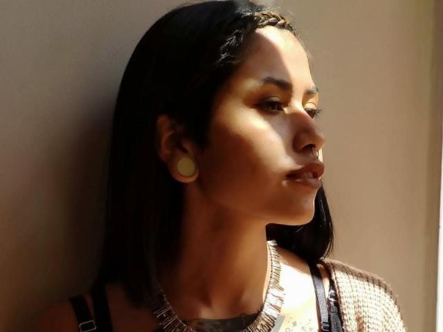 Photographie de NyxVega, avec des cheveux foncés, une peau brun clair et une expression sérieuse. La lumière du soleil illumine son visage, créant des ombres. Elle porte un haut en maille et de grandes boucles d'oreilles rondes. Regard incroyablement intense..