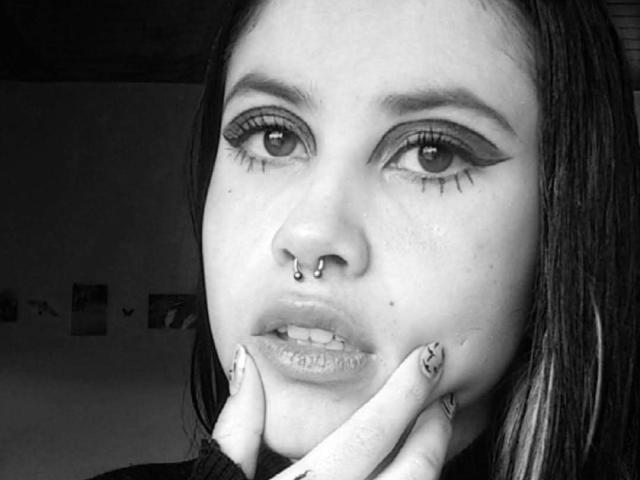 Fotografía en blanco y negro de NyxVega, primer plano, mano sujetando la barbilla, delineador de ojos marcado, piercing en la nariz, aro en el labio, cabello oscuro. fondo sombrío, alto contraste, mirada intensa, sugerente, ambiente provocador..