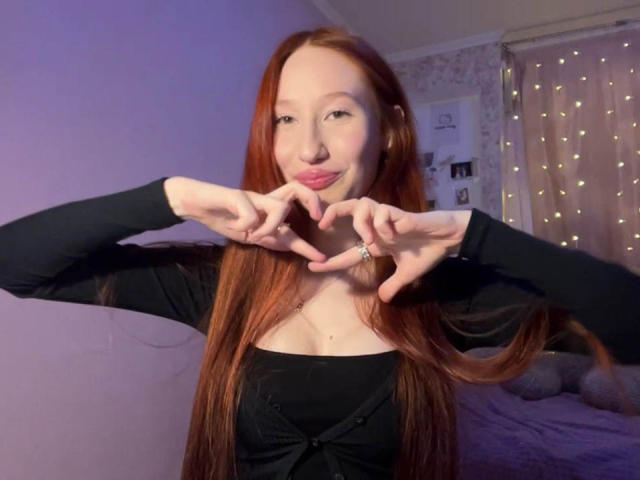 TrinhDancer, una giovane donna con lunghi capelli rossi, forma un cuore con le dita, indossando una maglietta nera, in un ambiente scarsamente illuminato. stanza con luci a stringa sullo sfondo. La sua espressione è leggermente seducente..