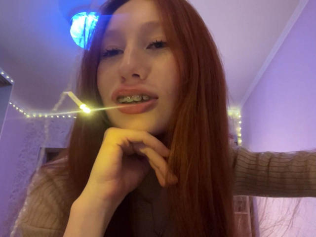 Nahaufnahme von TrinhDancer mit langen roten Haaren, die eine leuchtende Lichterkette anbeißt. Sie schaut mit einem leichten Lächeln in die Kamera und trägt einen braunen, gerippten Pullover. Verdammt heiß..