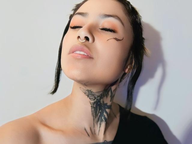 Fotografía de primer plano de NyxVega, una mujer de piel clara con cabello negro, ojos cerrados, un piercing en la nariz y un tatuaje tribal negro en el cuello. Ella tiene una blusa negra, una expresión desafiante y reflejos suaves..