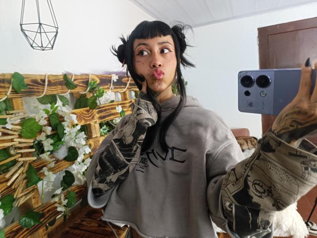 NyxVega takes a selfie, holding a smartphone with tattooed hands. Wearing a gray sweatshirt, black bangs, and messy hair, with a pouty expression. Wicker and flower arrangement in background.