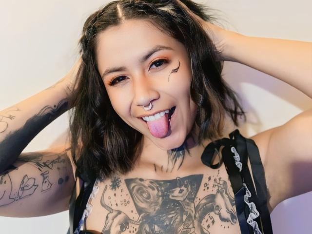 NyxVega met zwart haar, die haar tong uitsteekt, een neusring en tatoeages op haar borst en armen. Zwart tanktop aan, handen in het haar. Heldere, witte achtergrond. Heel speels uitgedrukt..