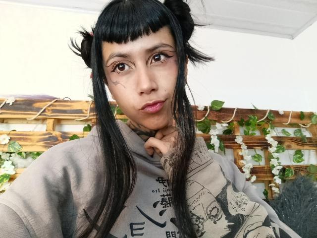 NyxVega, com cabelo preto e liso em dois coques, vestindo um moletom cinza com estampas, olhando diretamente para a câmera com uma expressão ligeiramente curiosa, com a mão tocando o queixo. Fundo externo claro..