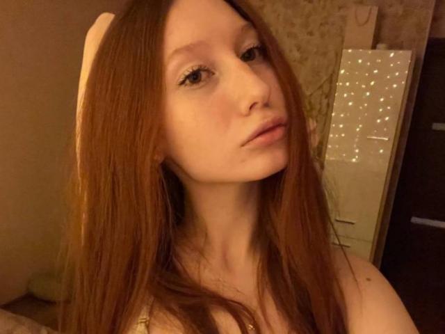 Autofoto de primer plano de TrinhDancer, una mujer de piel clara con cabello largo y liso de color marrón, mirando directamente a la cámara. Una expresión ligeramente seductora. Iluminación cálida, luces decorativas en el fondo.