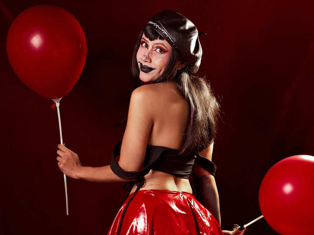 NyxVega, with black lipstick and long black hair, holds a red balloon in her right hand, wearing a black strapless top and shiny red skirt, looking back provocatively. Two red balloons in background.
