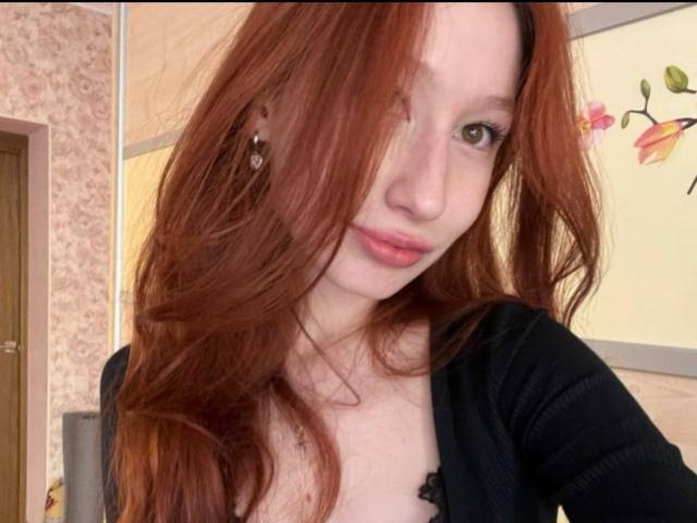 A TrinhDancer tem cabelos longos e ruivos que cobrem um olho, pele clara e um sorriso discreto. Ela veste uma blusa preta, que revela um pouco o colo. Seus olhos castanhos olham diretamente para a câmera..