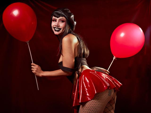 NyxVega, een aantrekkelijke vrouw met zwarte lippenstift en lang zwart haar, die twee rode ballonnen vasthoudt, gekleed in een rode leren rok, visnetkousen, zwarte top en zwarte hoofdband, met een verleidelijke blik over de schouder kijkend..