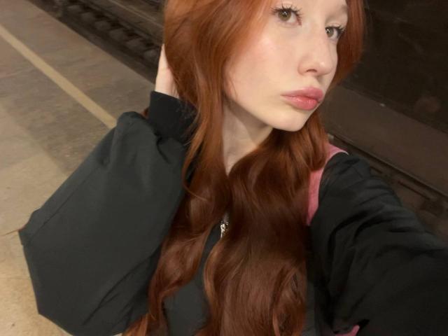 Close-up photo of TrinhDancer, a white woman with long, wavy red hair, wearing a black top, posing with her hand in her hair, pouty expression, slight shadow on her face.