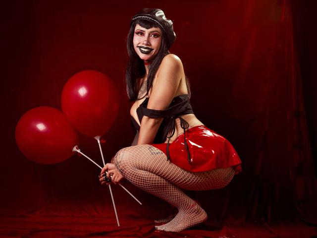 NyxVega squats with red balloons, wearing a black top, red latex skirt, fishnet stockings, and a black hat. She has black lipstick, holding sticks, against a dark red background.