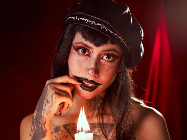 Gros plan de NyxVega avec des cheveux noirs, un maquillage gothique et des tatouages, portant un chapeau noir, regardant de manière séduisante une bougie allumée. éclairage rouge, regard intense, rouge à lèvres foncé, doigt près de la bouche.