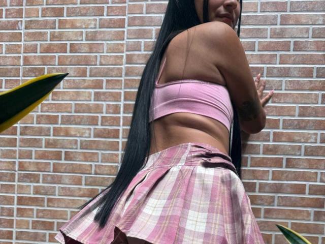 AnnieBlakke, con cabello negro largo, vestida con un sujetador rosa y una falda de cuadros, apoyada contra una pared de ladrillo, mostrando su espalda. pose coqueta, ligera curvatura en la espalda, tensión en su postura.