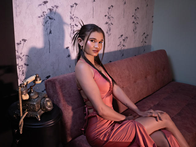 JennaDerose, una donna asiatica attraente con lunghi capelli neri, è seduta su un divano di velluto rosa, indossando un abito rosa aderente e lucido. Le ombre mettono in risalto la sua figura sinuosa, creando una tensione sessuale..