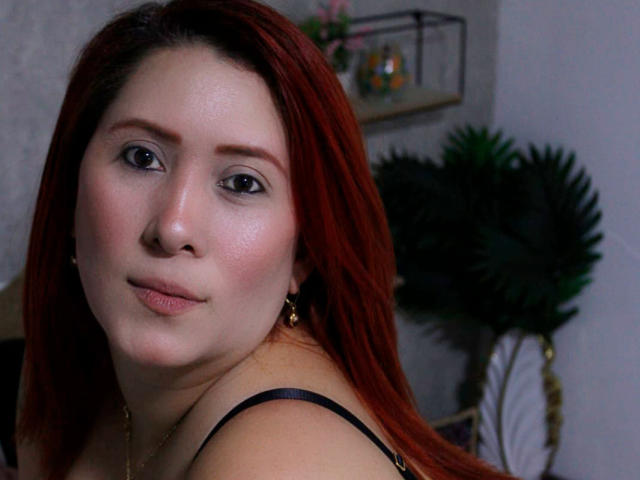 Primer plano de AfroditaMartinez, una mujer de piel clara y cabello rojo, que lleva un top negro con tirantes y mira directamente a la cámara con una sonrisa sutil, una luz suave que resalta sus mejillas sonrojadas. El fondo incluye una planta y una estantería..