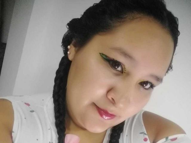 Close-up selfie of CattaleyaBohorquez, an Asian woman with dark braids, green eyeliner, and pink lipstick, looking directly at the camera with a slight smile. Visible chest and white floral top.