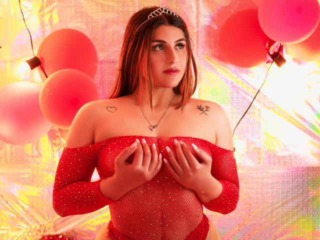 SaraWestt è in piedi davanti a dei palloncini rossi, indossa una top rossa in rete con spalline scoperte e tiene le mani sul suo piccolo seni. Ha una tiara, tatuaggi sulle spalle e un'espressione seducente. Sfondo luminoso e colorato.