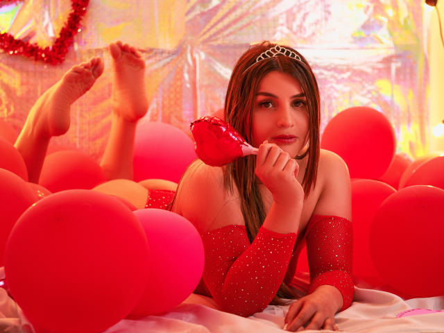 SaraWestt mit dunkelbraunen Haaren, die ein rotes, glitzerndes Outfit trägt und inmitten von roten, herzförmigen Ballons liegt, lutscht an einem roten lutscher, gebeugte Beine, Füße angezogen, intensiver Blick, lebhaftes Rot- und Orangetöniges Licht..