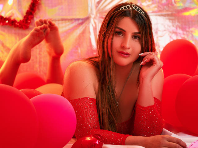 SaraWestt lies in a bed of red balloons, wearing a sparkly red off-shoulder top and a tiara. Her long braids fall over her shoulders, holding a phone to her ear. Fucking sexy.