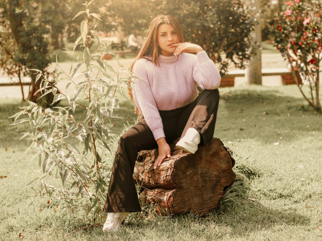 SaraWestt, a young woman with long brown hair, sits on a tree stump in a sunlit park. She wears a white knit sweater and black pants, leaning forward with a seductive smile.