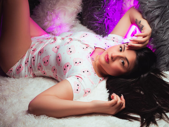 SaraWestt lies on a white, furry surface, wearing a white, short-sleeve top with pink skull patterns. Her hand touches her forehead, glowing pink light behind her. Fucking sexy, relaxed pose.
