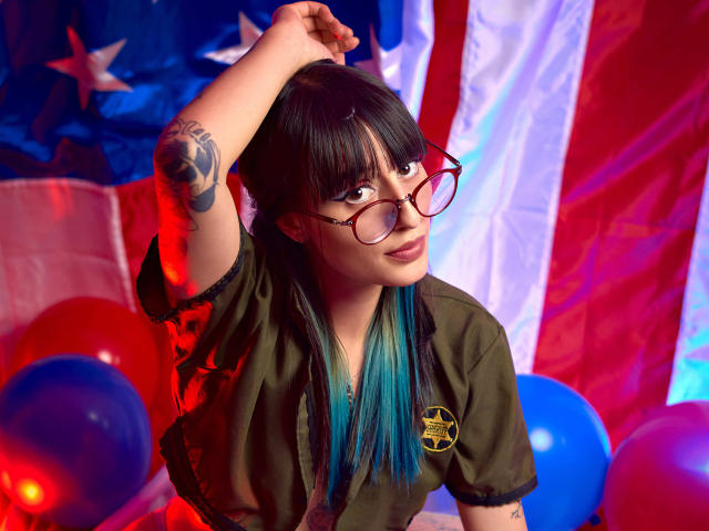 TatsumiLee with black and blue hair, wearing glasses and a green shirt, raises arm, revealing tattoo. American flag in background, red and blue balloons. Sexy, confident pose.