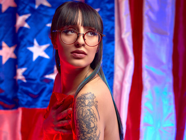 TatsumiLee met recht zwart haar, een pony en ronde brillen, gekleed in een rode jurk, met een hand op de borst, een getatoeëerde schouder. tegen Amerikaanse vlaggen. Rode en blauwe verlichting, sensueel, intense blik.