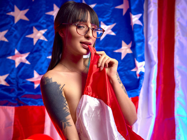 TatsumiLee, Asian with long black hair and glasses, holding red fabric against an American flag backdrop. She's topless, showing a tattoo on her left arm. Seductive, licking her finger.