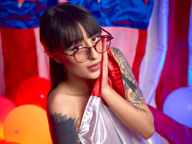 TatsumiLee, a tattooed woman with black hair and red glasses, holds a red ribbon, wearing a white strapless top. Bright colors, sexy, seductive pose, vivid tattoos, intense gaze.