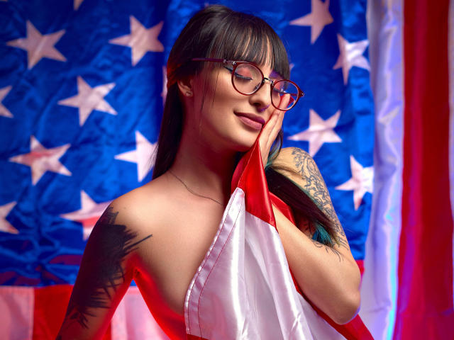 TatsumiLee, Asian woman with long black hair and glasses, holding a red and white flag, tattoos on shoulder, sexy, patriotic, stars and stripes background, vibrant colors, confident pose.