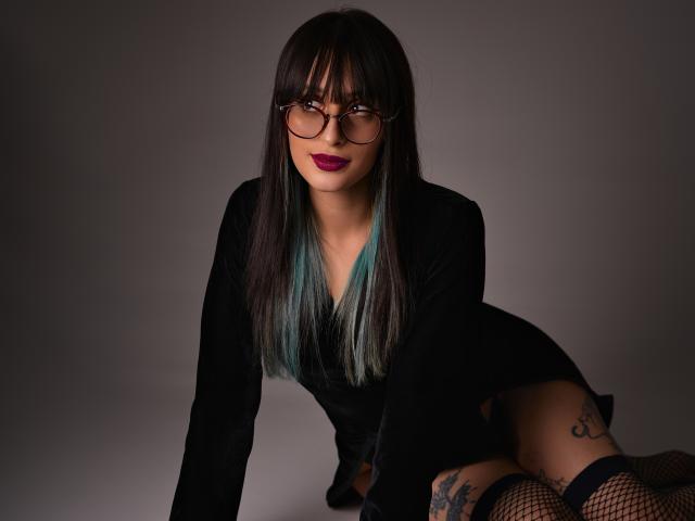 TatsumiLee, met zwart, steil haar, een bril en rode lippenstift, hurkt suggestief in een zwart colbert, met een net van visdraad. kousen, tatoeages op de dij, intense blik, donkere belichting, seksuele spanning, verleidelijke pose.