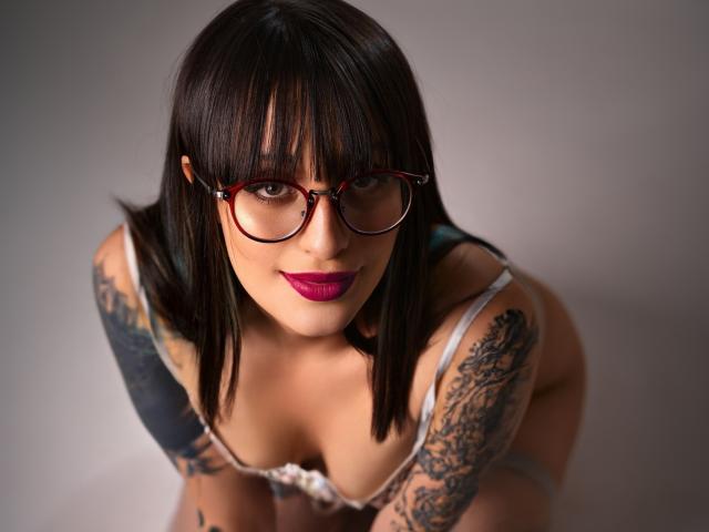 TatsumiLee, avec ses cheveux noirs et lisses, ses lunettes rouges et son rouge à lèvres foncé, penchée en avant, regardant vers le haut, portant un soutien-gorge blanc. qui révèle la poitrine, des tatouages sur les bras, un regard intense, une pose provocante..