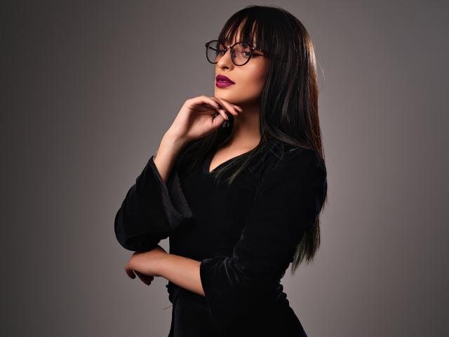 TatsumiLee, avec ses longs cheveux noirs, portant des lunettes noires et une robe noire, pose la main près du menton, avec un rouge à lèvres foncé. Expression sensuelle, éclairage de studio doux, fond gris, ambiance sexy..