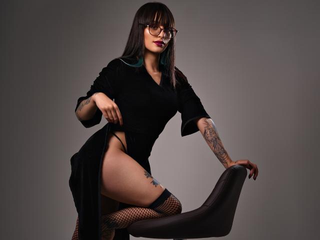 TatsumiLee, vestida con un vestido negro, medias de rejilla y gafas, se arrodilla con una pierna levantada, exponiendo su muslo. Tatuajes en el brazo, cabello oscuro, lápiz labial rojo, mirada intensa, pose provocativa, fondo gris..
