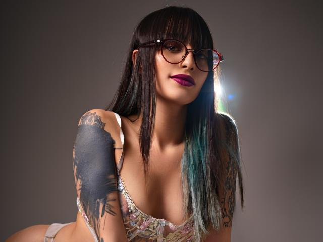 TatsumiLee, a sexy woman with long black and blue hair, wearing glasses and a low-cut bra, exposing her ample cleavage. She has a tattoo on her left shoulder. Bright light behind her.